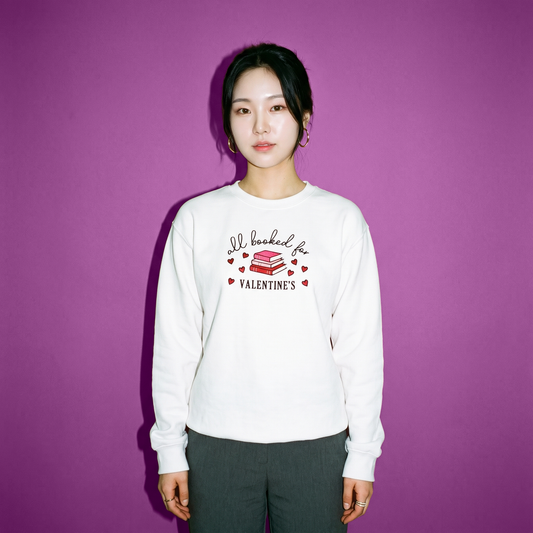 A woman stands against a solid purple background wearing a white long-sleeve crewneck sweatshirt with a graphic that reads 'All booked for VALENTINES' with a stack of red books and small red hearts. She is also wearing dark gray pants.